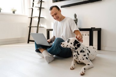 Genç, orta yaşlı bir adam evde köpeğiyle yerde otururken dizüstü bilgisayarda çalışıyor.