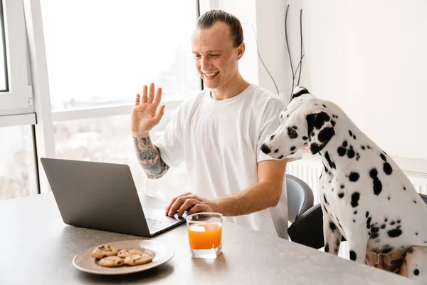 Smiling mid aged man working on laptop computer while sitting at the kitchen wit his dog, having breakfast, waving hand on a video call
