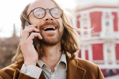 Gözlüklü kızıl adam caddede cep telefonuyla konuşurken gülüyor.