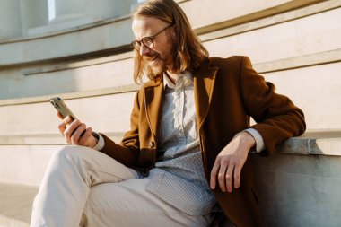 Kızıl adam merdivenlerde otururken gülümsüyor ve cep telefonu kullanıyor.