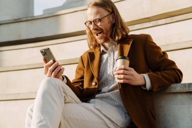 Kızıl adam merdivenlerde otururken cep telefonu kullanıyor ve kahve içiyor.