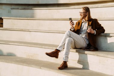 Kızıl adam merdivenlerde otururken cep telefonu kullanıyor ve kahve içiyor.