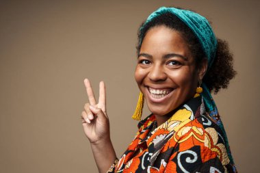 Saçbandı takmış, gülümseyen ve barış işareti gösteren genç siyahi kadın gri arka planda izole edilmiş.