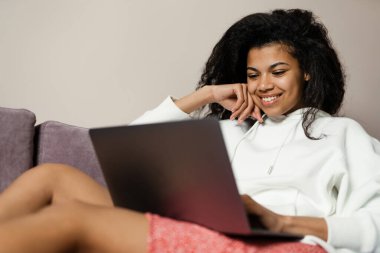 Genç siyahi kadın evdeki koltukta otururken dizüstü bilgisayar kullanıyor.