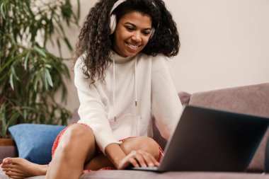 Kulaklıklı genç siyahi kadın evdeki kanepede otururken dizüstü bilgisayar kullanıyor.