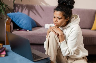 Evdeki kanepede otururken dizüstü bilgisayar kullanan Afrikalı genç bir kadın.