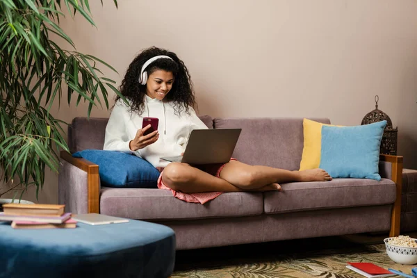 Young black woman in headphones using laptop and cellphone while sitting on sofa at home
