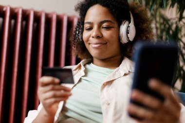 Evde cep telefonu ve kredi kartı kullanan kulaklıklı, gülümseyen genç bir kadın.