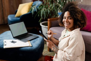 Genç siyahi kadın evde dizüstü bilgisayarla çalışırken cep telefonu kullanıyor.