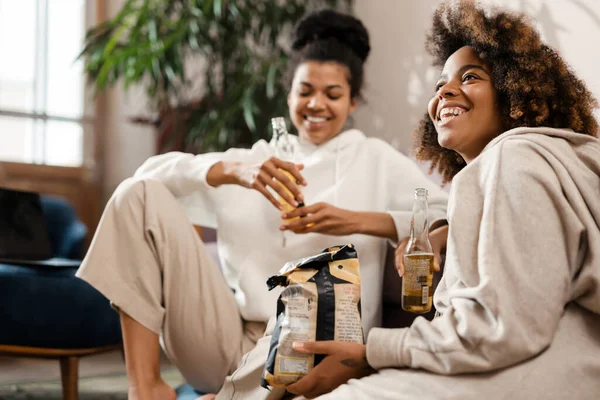 Две Счастливые Африканские Подружки Сидят Диване Пьют Пиво Поедая Чипсы — стоковое фото