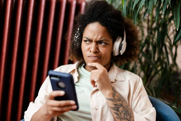 Νεαρή Μαύρη Δυσαρεστημένη Γυναίκα Ακουστικά Που Χρησιμοποιεί Κινητό Στο Σπίτι — Φωτογραφία Αρχείου