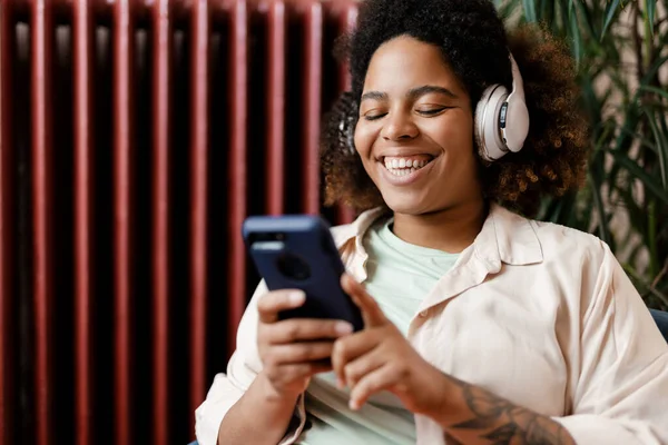 自宅で携帯電話を使用してヘッドフォンで若い黒人の笑顔の女性 — ストック写真