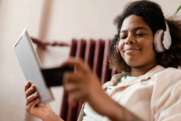 在家里使用平板电脑和信用卡的戴耳机的年轻黑人妇女 — 图库照片