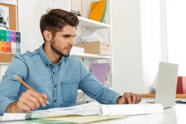 Genç beyaz adam dizüstü bilgisayarla çalışıyor ve ofiste çizimler yapıyor.