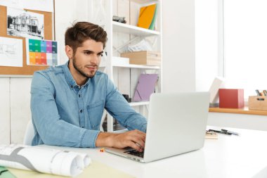 Genç beyaz adam dizüstü bilgisayarla çalışıyor ve ofiste çizimler yapıyor.