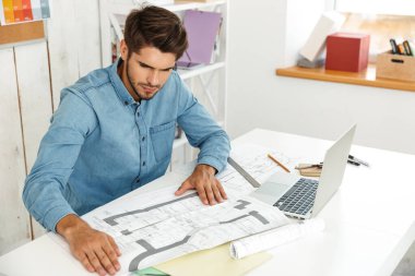 Genç beyaz adam dizüstü bilgisayarla çalışıyor ve ofiste çizimler yapıyor.