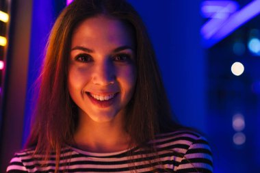 Çizgili kıyafetler giyen genç beyaz kadın gülümsüyor ve kapalı alanda kameraya bakıyor.