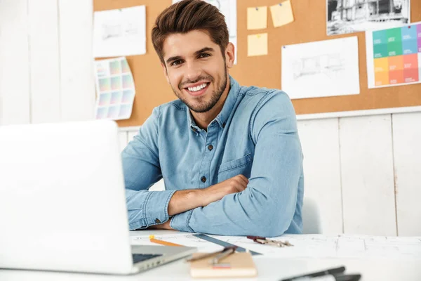 Νεαρός Λευκός Άνδρας Χαμογελά Ενώ Εργάζεται Laptop Και Σχέδια Εσωτερικό — Φωτογραφία Αρχείου