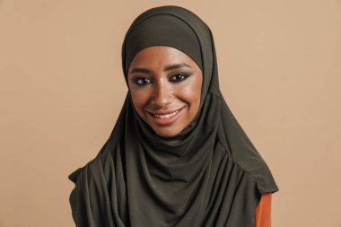 Başörtülü genç Müslüman kadın gülümsüyor ve bej arka planda izole edilmiş kameraya bakıyor.