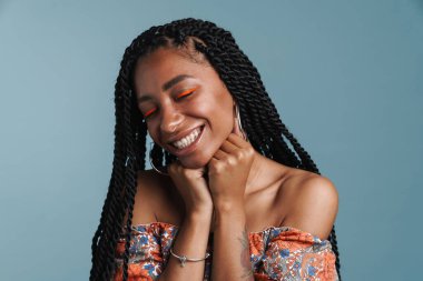 Makyajlı, gülümseyen ve kameraya poz veren genç siyah kadın mavi arka planda izole edilmiş.