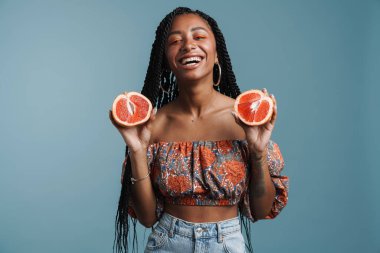 Mavi arka planda izole edilmiş greyfurt gösterirken makyajlı genç siyah kadın gülümsüyor.
