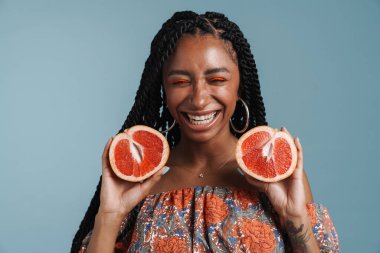 Mavi arka planda izole edilmiş greyfurt gösterirken makyajlı genç bir zenci kadın gülüyor.