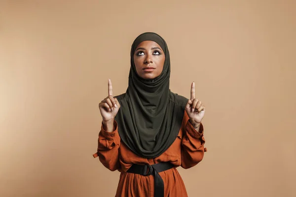 Молода Мусульманка Хіджабі Дивиться Вказує Пальці Вгору Ізольовано Бежевому Фоні — стокове фото