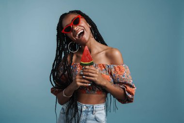 Güneş gözlüklü genç siyah bir kadın mavi arka planda lolipopla poz verirken gülüyor.