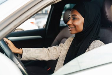 Siyah Müslüman kadın araba kullanırken başörtüsü takıyor gülümsüyor.
