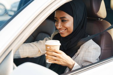 Siyah Müslüman kadın başörtüsü takıyor. Araba kullanırken kahve içiyor.