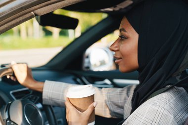 Siyah Müslüman kadın başörtüsü takıyor. Araba kullanırken kahve içiyor.