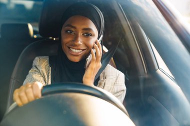 Siyah Müslüman kadın başörtüsü takıyor. Araba kullanırken cep telefonuyla konuşuyor.
