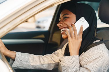 Siyah Müslüman kadın başörtüsü takıyor. Araba kullanırken cep telefonuyla konuşuyor.
