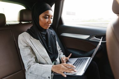 Siyah Müslüman kadın başörtüsü takıyor. Arabada dizüstü bilgisayarla çalışıyor.