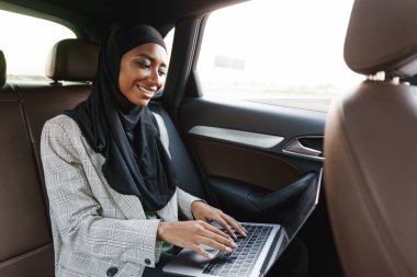 Siyah Müslüman kadın başörtüsü takıyor. Arabada dizüstü bilgisayarla çalışıyor.