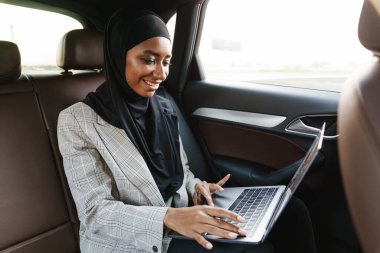Siyah Müslüman kadın başörtüsü takıyor. Arabada dizüstü bilgisayarla çalışıyor.