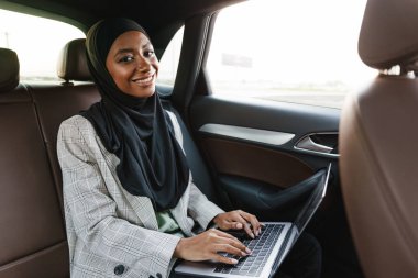 Siyah Müslüman kadın başörtüsü takıyor. Arabada dizüstü bilgisayarla çalışıyor.