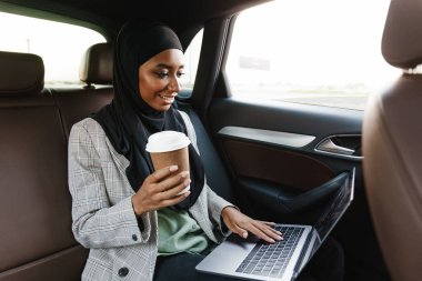 Başörtüsü takan siyah Müslüman kadın laptopla çalışıyor ve arabada kahve içiyor.