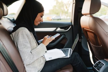 Arabadayken cep telefonu ve laptopla çalışan başörtüsü takan siyahi bir kadın.