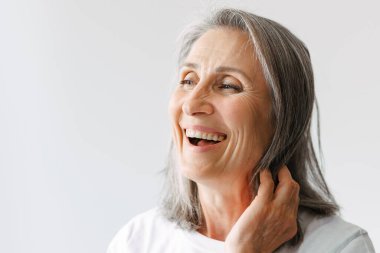 Gri tişörtlü, gülen ve beyaz arka planda tek başına kalan yaşlı bir kadın.