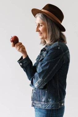 Beyaz arka planda izole edilmiş elmayla poz verirken şapkalı, gülümseyen gri yaşlı kadın.