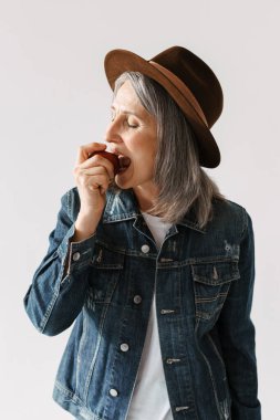 Şapkalı, elma yiyen, gözleri kapalı, beyaz arka planda yalnız, yaşlı bir kadın.