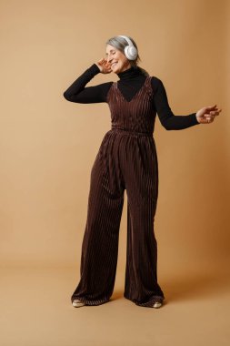 Bej arka planda izole edilmiş kulaklıklarla müzik dinlerken gülümseyen yaşlı gri kadın.