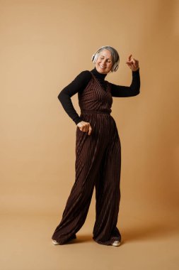 Bej arka planda izole edilmiş kulaklıklarla müzik dinlerken dans eden yaşlı gri kadın.