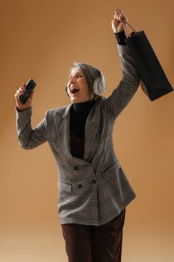 Gri ceketli, kulaklık ve cep telefonu kullanan, bej arka planda izole edilmiş yaşlı kadın.