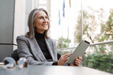 Açık kafede otururken tablet bilgisayar kullanan gri beyaz yaşlı kadın.