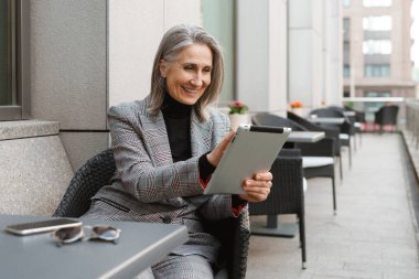 Açık kafede otururken tablet bilgisayar kullanan gri beyaz yaşlı kadın.