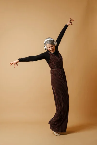灰色老年妇女一边听音乐一边跳舞 耳机与米色背景隔离 — 图库照片
