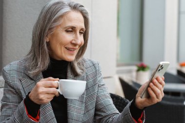 Gri beyaz yaşlı kadın cep telefonu kullanıyor ve açık kafede kahve içiyor.