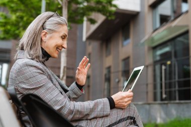 Açık havada bankta otururken tablet bilgisayar kullanan gri beyaz bir kadın.